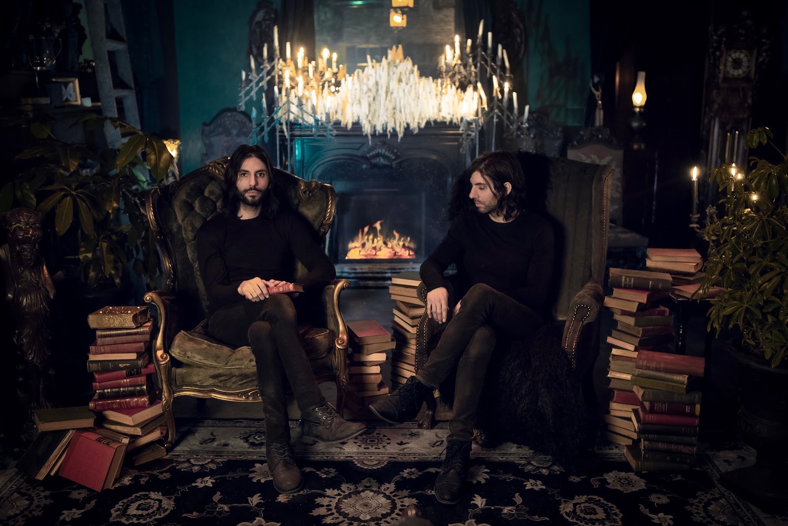 Twin brothers Ryan and Matthew Murray of Black Veil Studio seated in armchairs by a fire in their candlelit Salem shop, surrounded by stacks of antique books