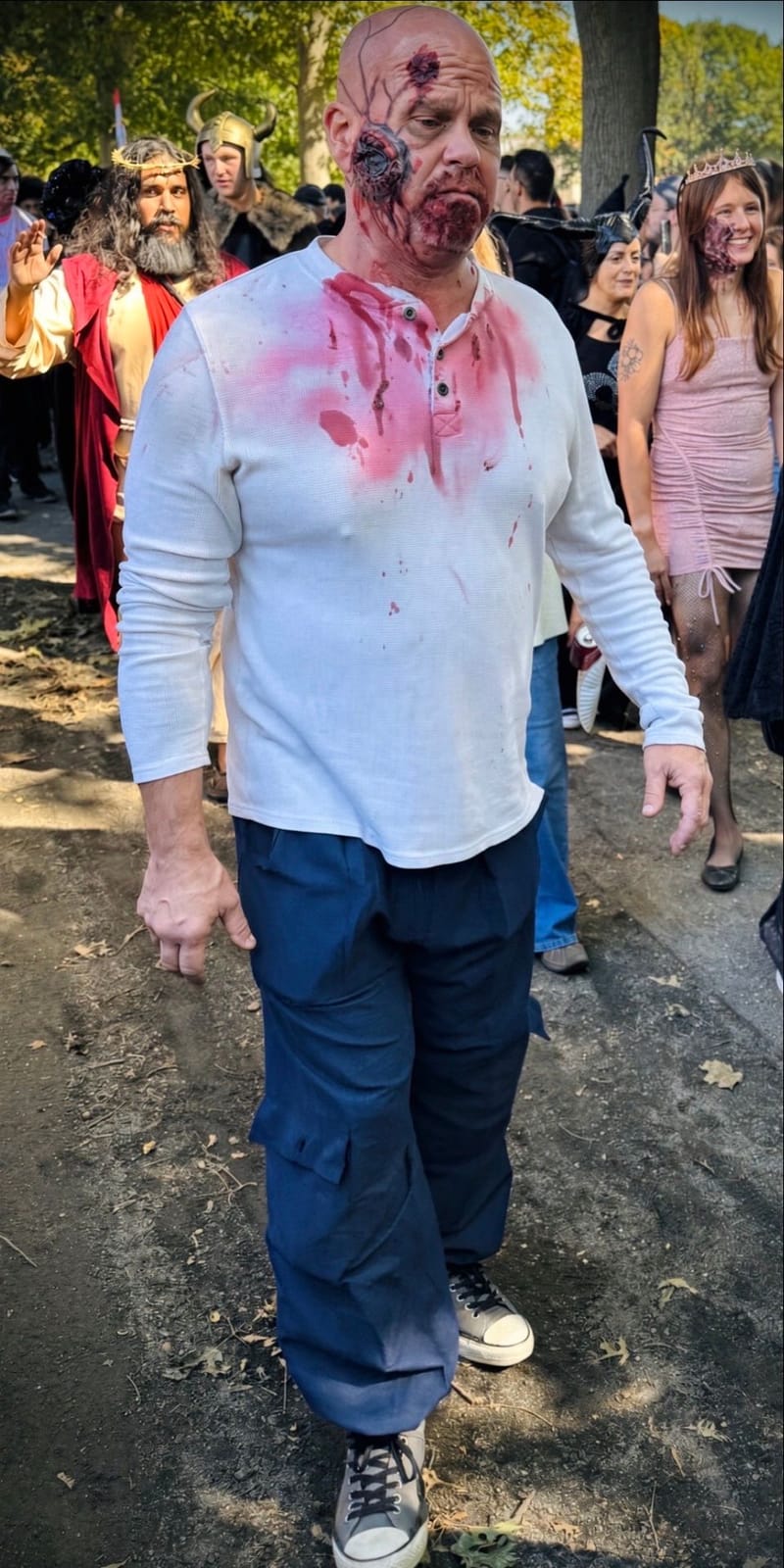 A bald man in zombie makeup with a gaping wound on his cheek and blood soaking his white henley, walking with other costumed zombies on Salem Common