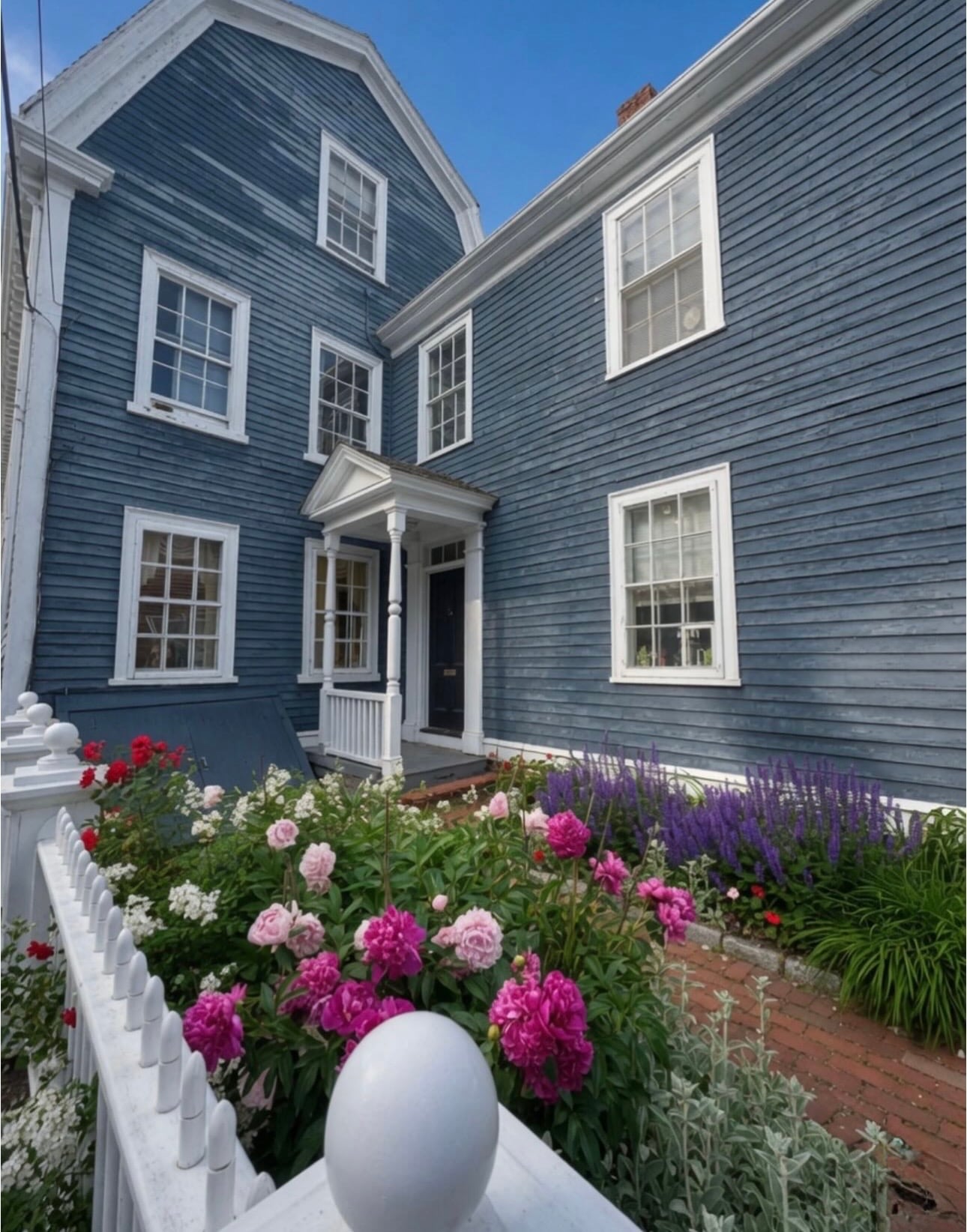 A slate-blue gambrel-roofed colonial house with a pedimented doorway and a front garden of pink peonies and purple salvia behind a white picket fence in Salem