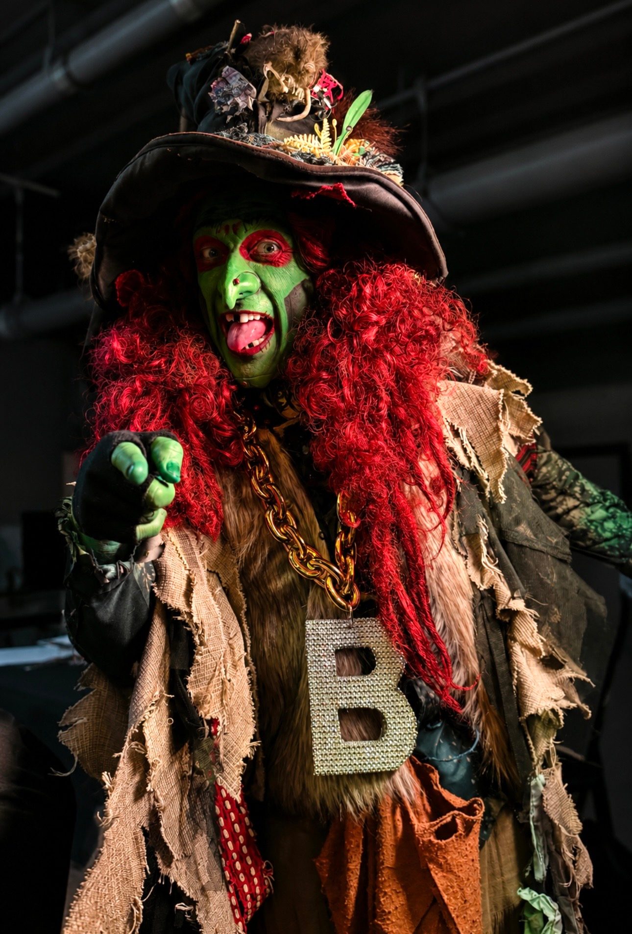 Borah the Witch, a green-faced performer with wild red hair and a jeweled 'B' pendant, pointing at the camera in Salem