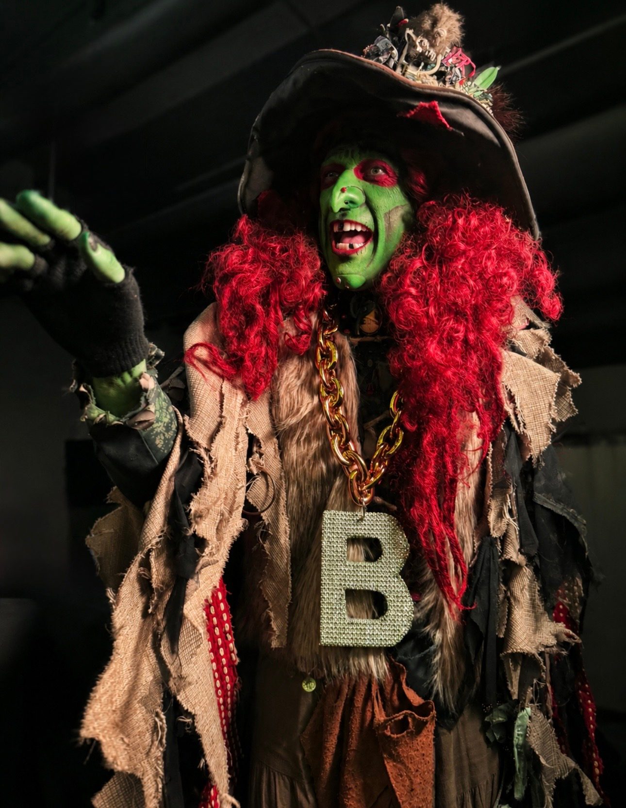 Borah the Witch, a green-faced Salem character performer with wild red hair, a battered hat, and a bejeweled 'B' chain
