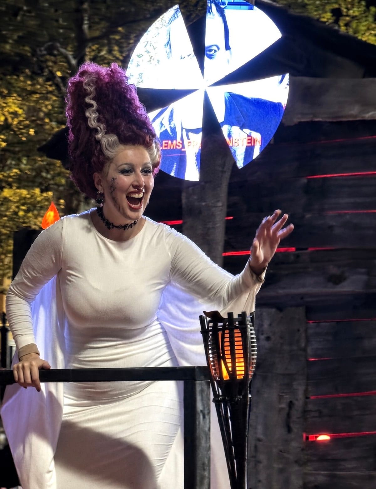 A woman as the Bride of Frankenstein, with a towering purple and white streaked beehive, white gown and stitched cheeks, waving from a float beside a lit torch and a Salem's Frankenstein screen