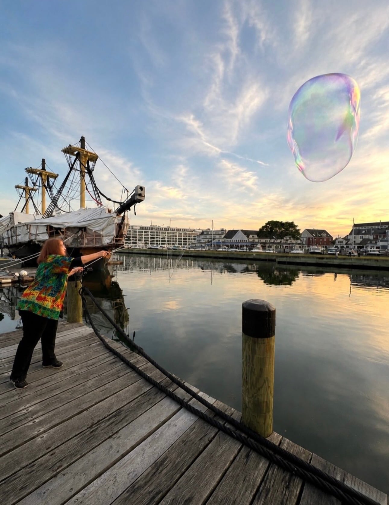 A street performer trailing a giant bubble along Derby Wharf beside the Friendship of Salem tall ship at golden hour