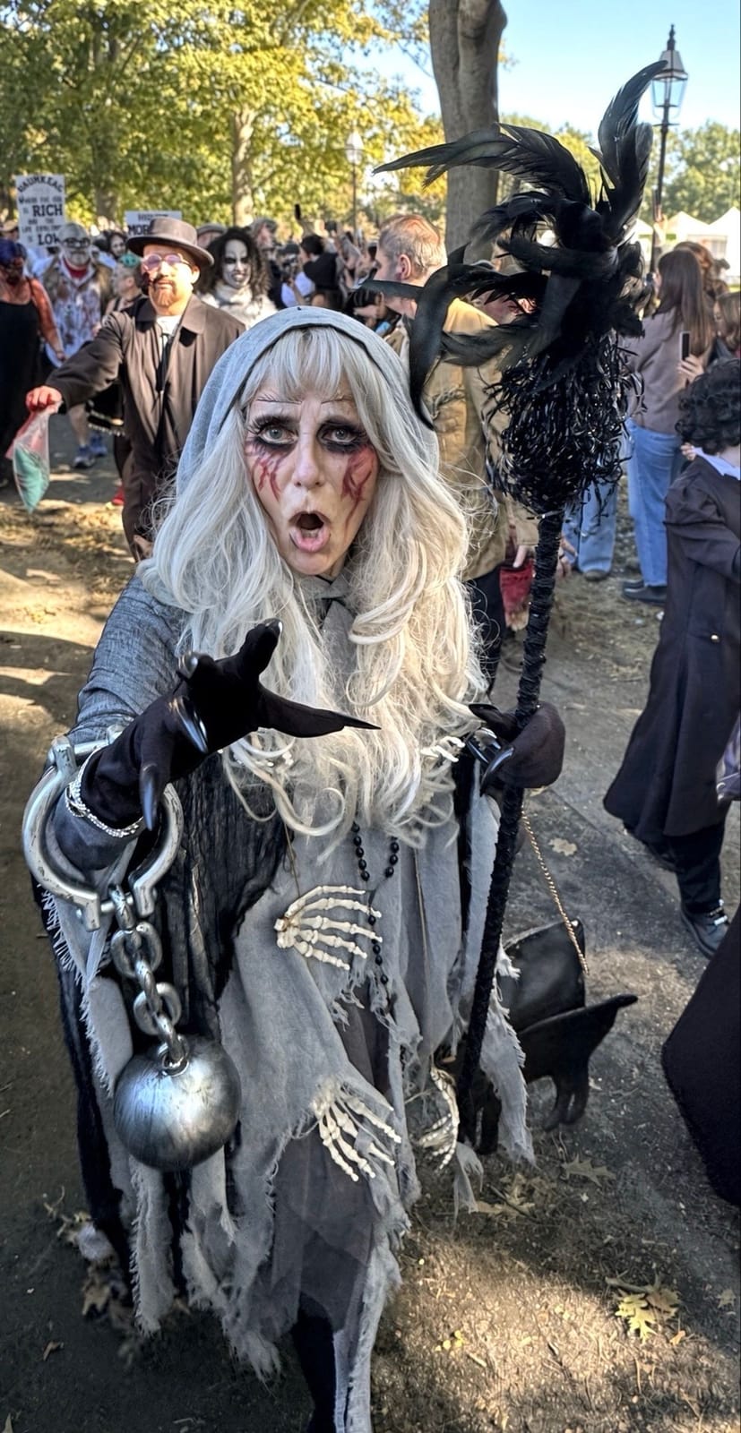 A woman with long white hair and smeared dark eye makeup in a gray tattered robe, dragging a ball and chain and holding a feathered staff, reaching toward the camera on Salem Common