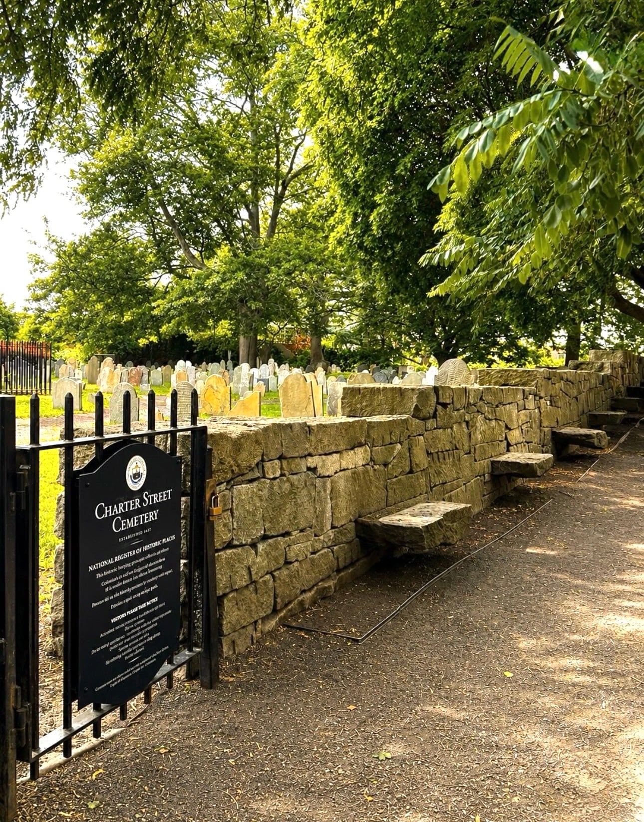 The gated entrance and dry stone wall of Charter Street Cemetery, Salem's oldest burying ground, with colonial slate headstones under summer trees