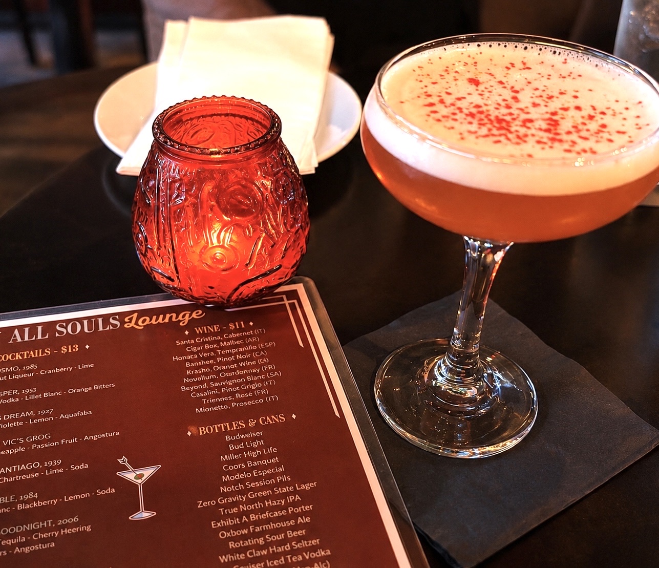 A coupe glass of pale orange cocktail topped with foam and red bitters, beside a red glass candle and the All Souls Lounge drinks menu