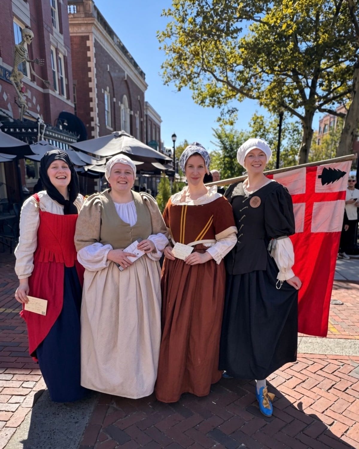 Four smiling women in 17th-century dress, with linen caps and coifs, bodices and long skirts in red, oatmeal, rust and black, standing on the Essex Street pedestrian mall holding paper tickets, beside a red flag with a white cross and a pine tree in the corner