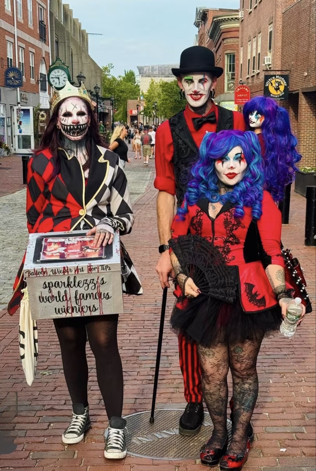 Three people in elaborate harlequin and circus costumes with full face paint on the brick Essex Street pedestrian mall in downtown Salem
