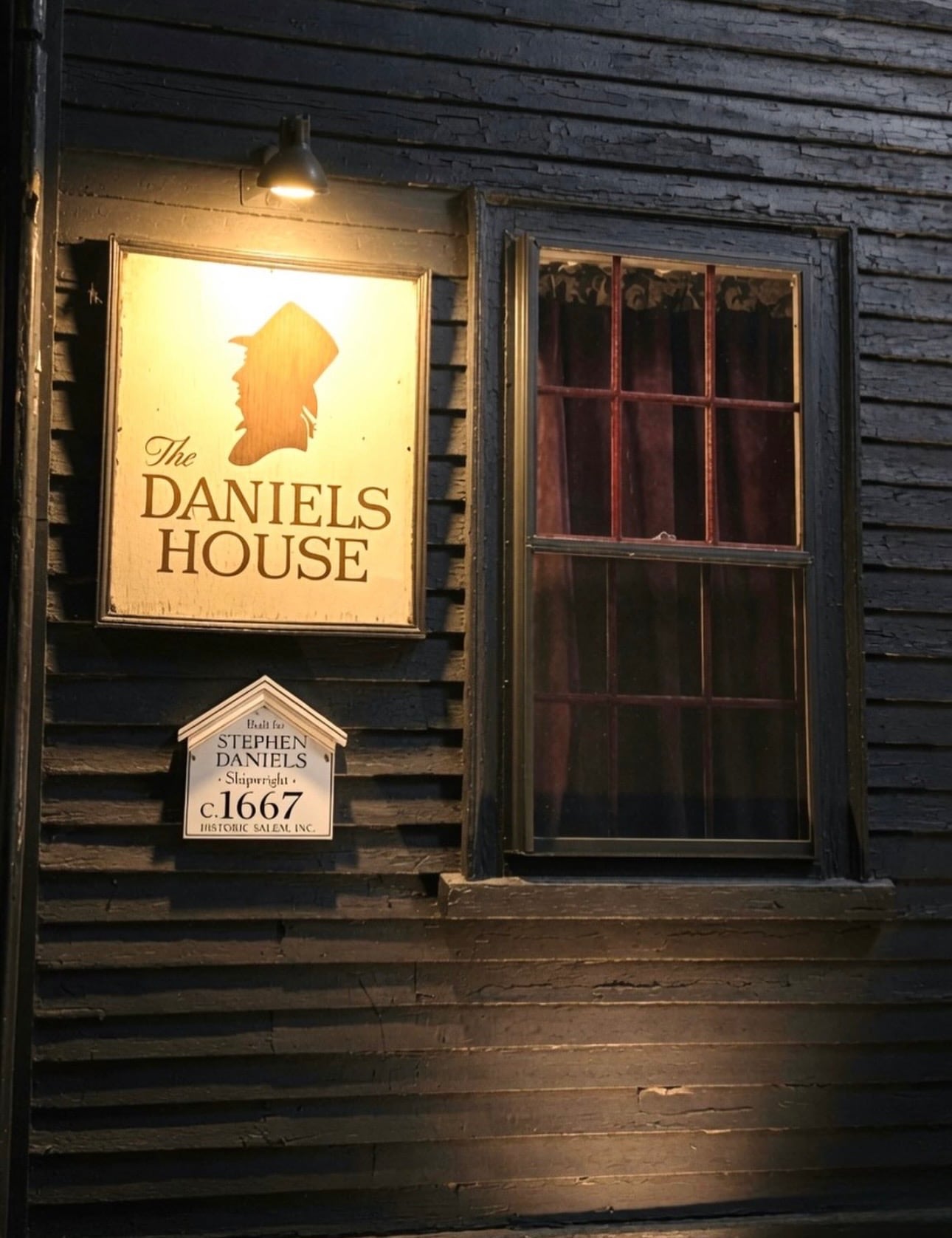 A glowing sign and 'c.1667' plaque on the dark clapboard wall of the Stephen Daniels House at night in Salem