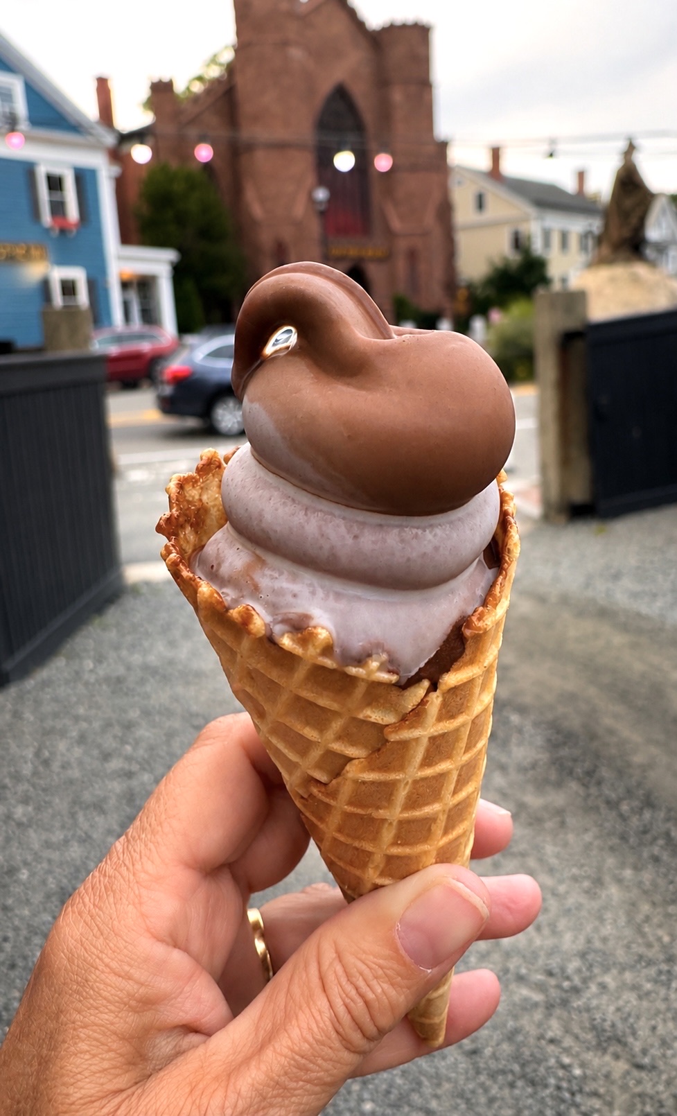A hand holding a chocolate-dipped soft serve cone in a waffle cone from Goodnight Fatty, with cafe string lights and a brownstone church behind on a Salem street
