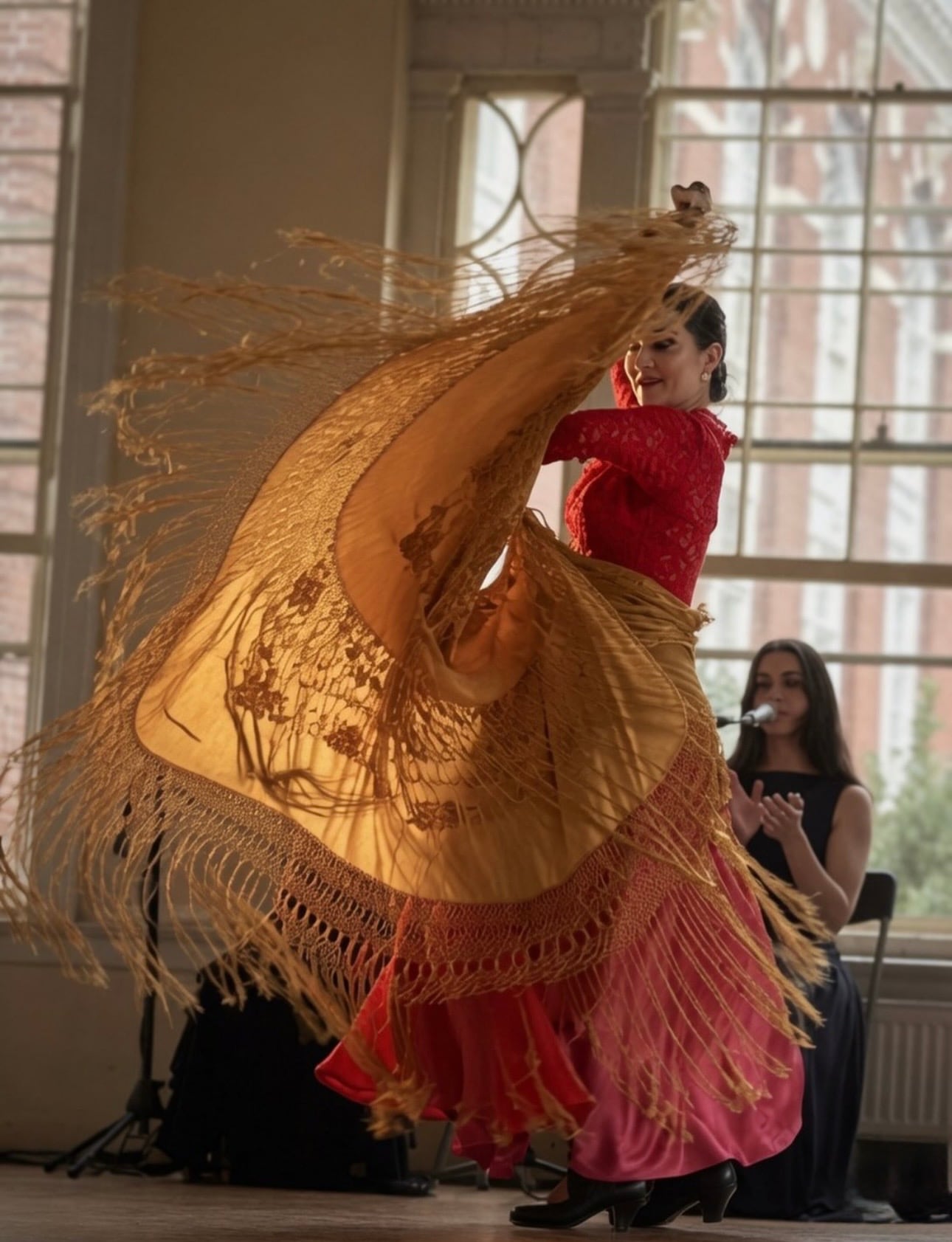 A flamenco dancer in a red top and golden fringed shawl mid-spin during a live performance by tall windows in Salem