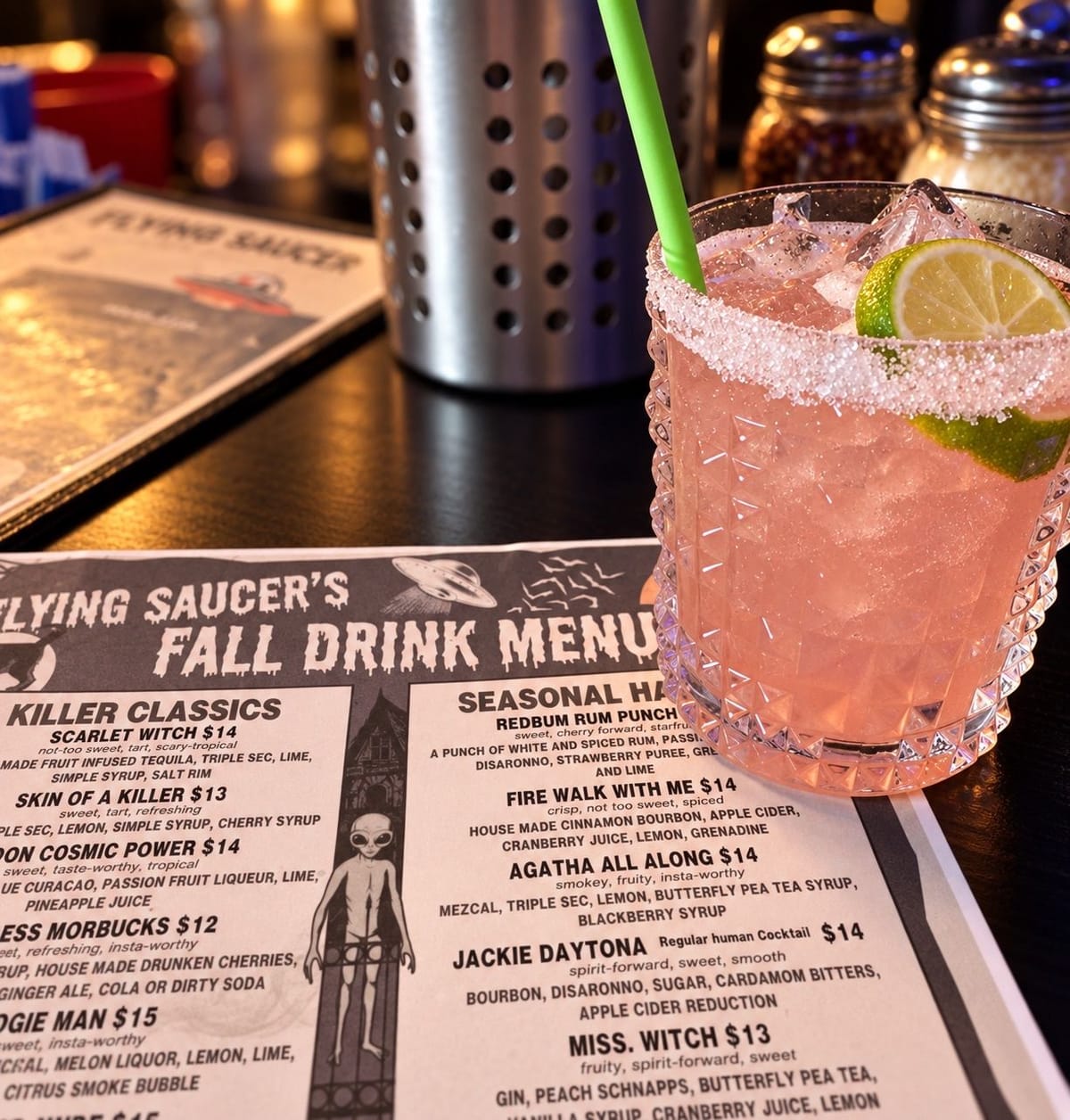 A pink cocktail with a sugared rim, a lime wheel and a green straw on a dark bar top, beside Flying Saucer Pizza Company's Fall Drink Menu listing Killer Classics and Seasonal Haunts cocktails