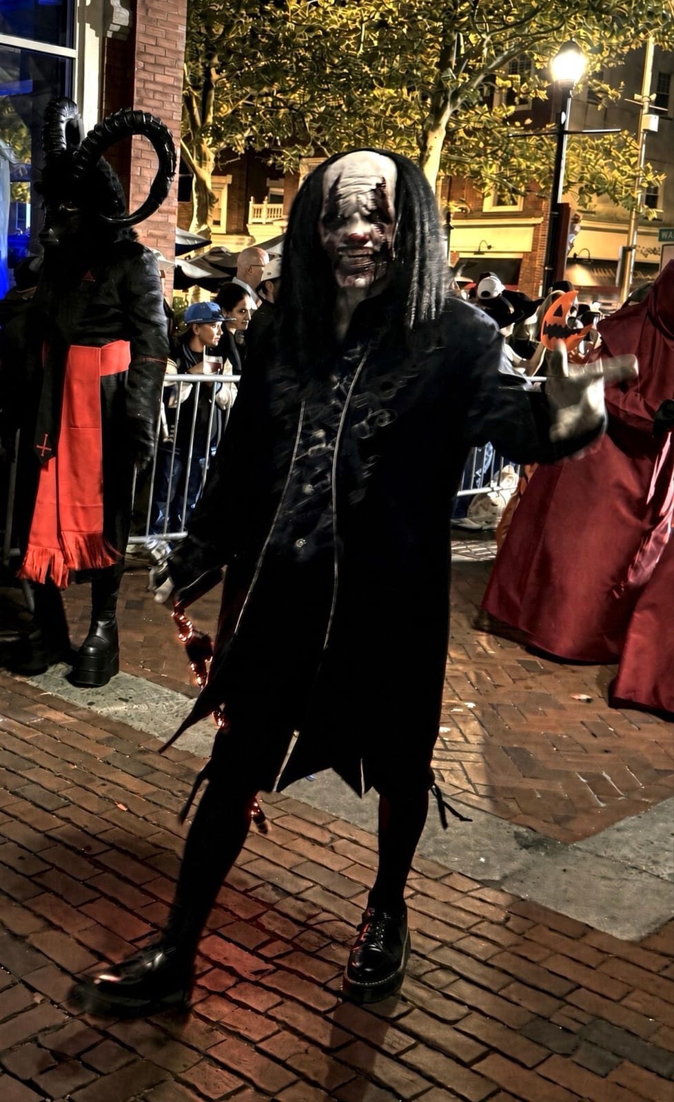 A grinning ghoul with long black hair and gray rotted makeup, reaching toward the camera in a black coat, with a hooded horned figure and a red-robed figure behind him on the parade route at night
