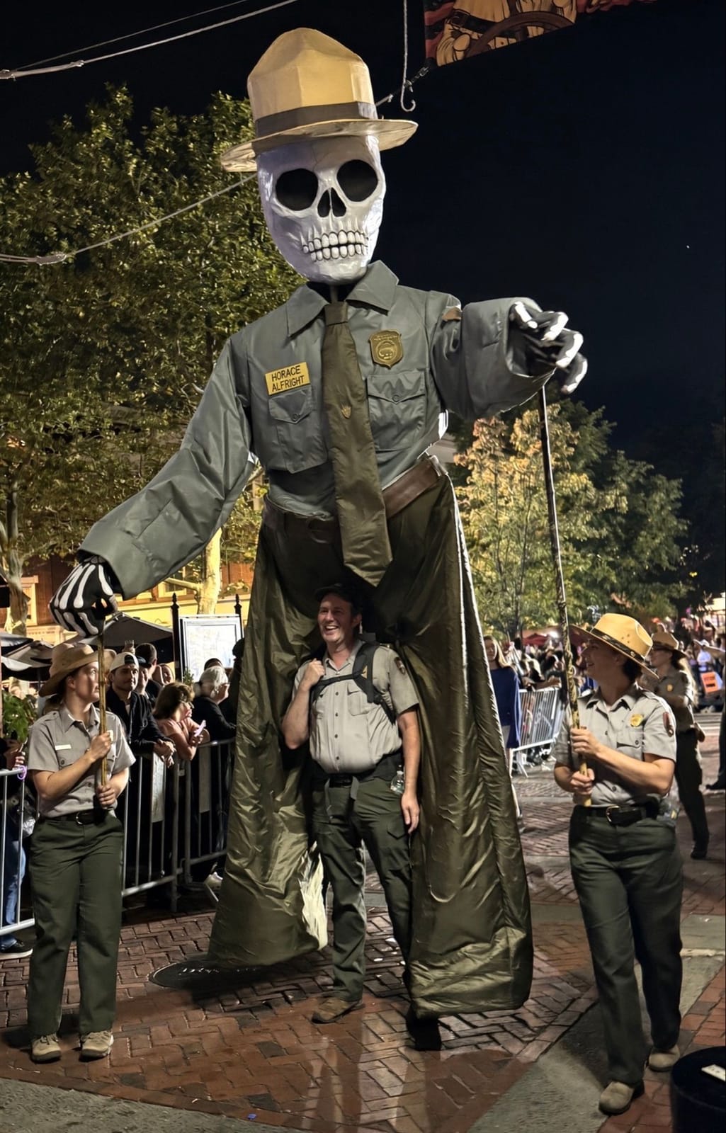A huge skeleton puppet in a National Park Service ranger uniform, flat hat and tie, carried by a ranger walking inside it, flanked by two uniformed park rangers at the Grand Parade at night
