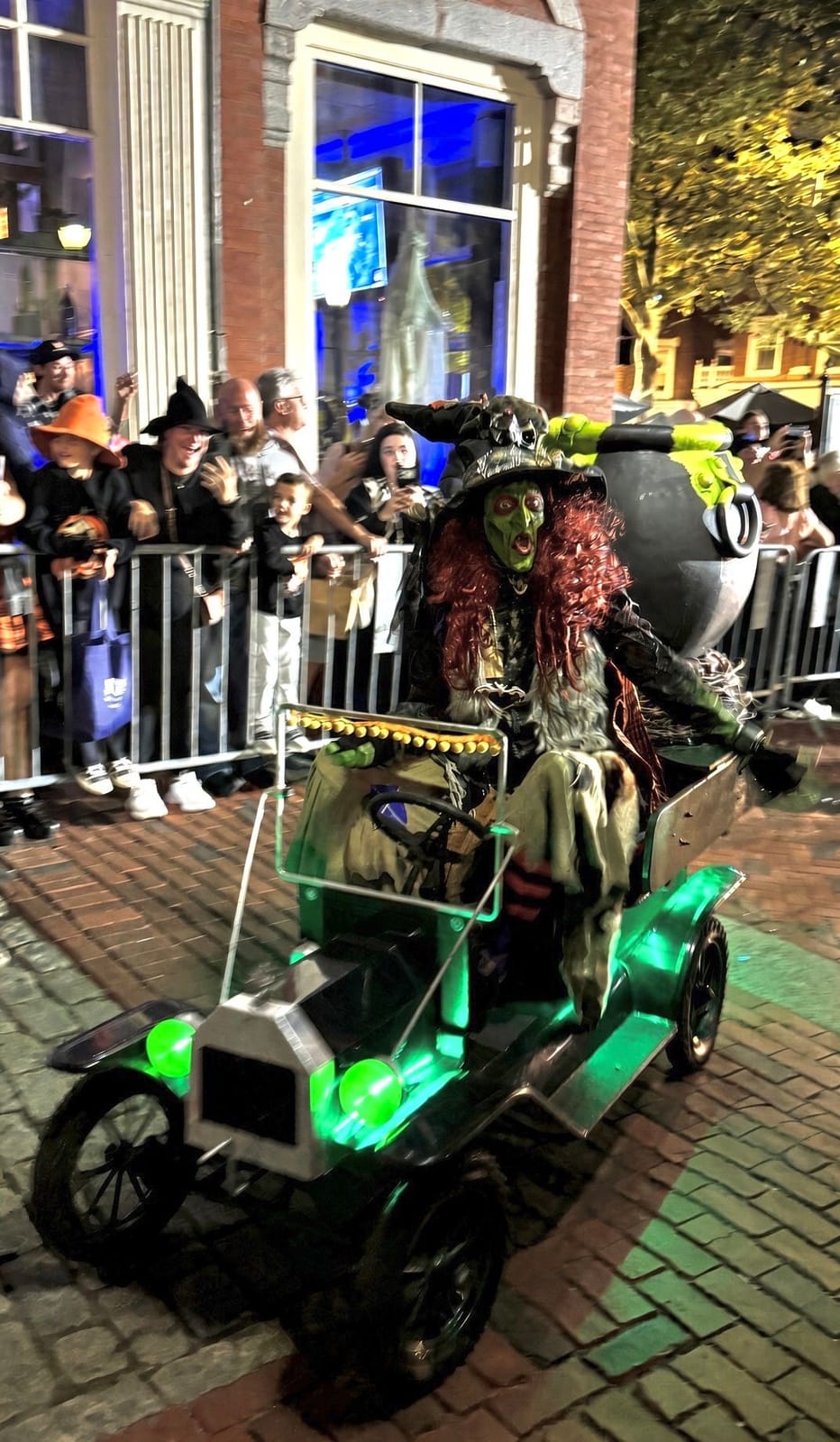 A green-faced witch with long red hair and a pointed hat driving a small antique-style car carrying a black cauldron with green ooze, lit green underneath, past the crowd behind the parade barricades at night