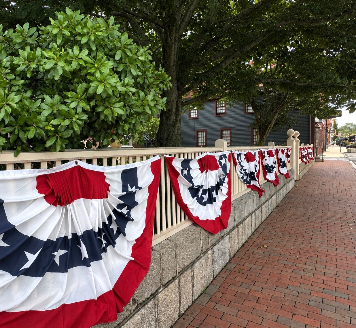Red-white-and-blue bunting draped along a white fence on a brick sidewalk in Salem, with a historic dark clapboard building behind