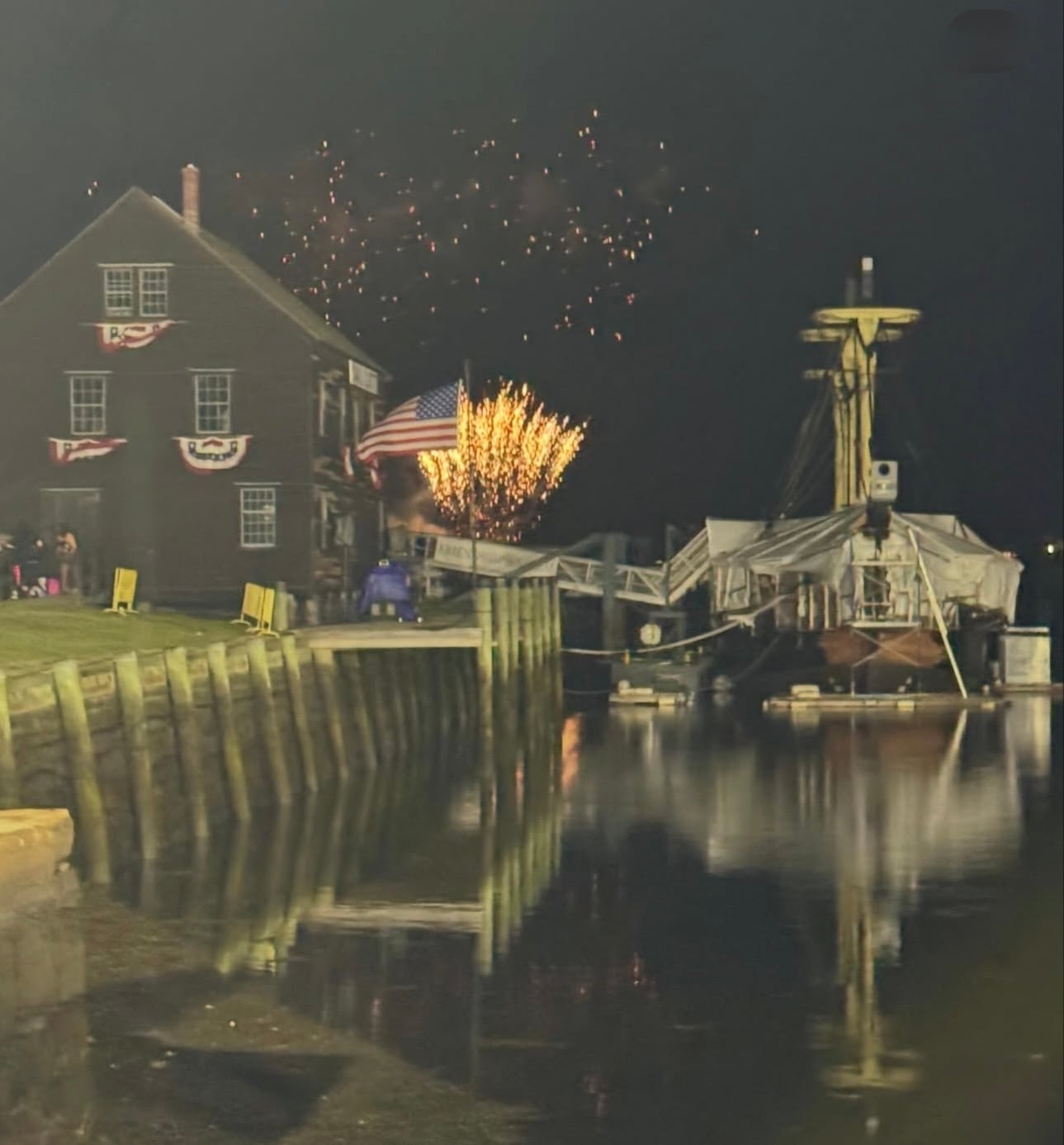 Fireworks over the Salem waterfront at night beside a bunting-draped warehouse and a tall ship at Derby Wharf