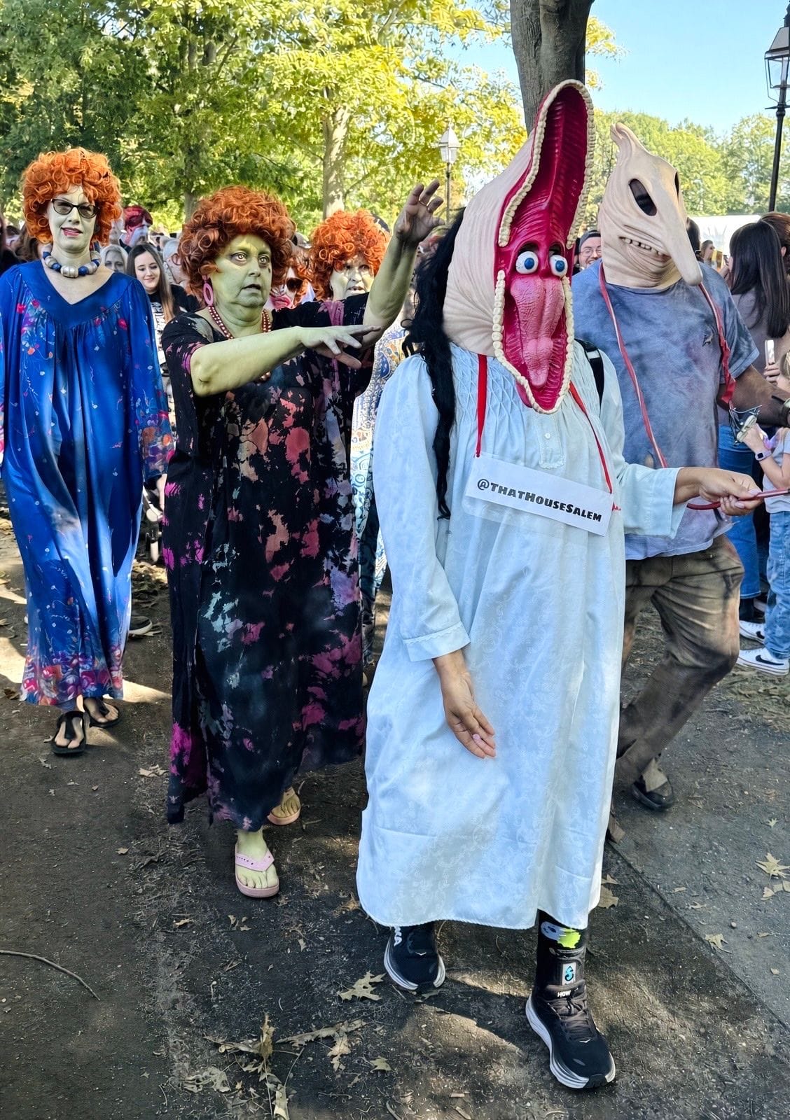 Three green-faced zombies in curly red wigs and bright caftans dressed as Mrs. Roper, walking behind two people in Beetlejuice's Maitland stretched-face scare masks, one with a sign reading @ThatHouseSalem