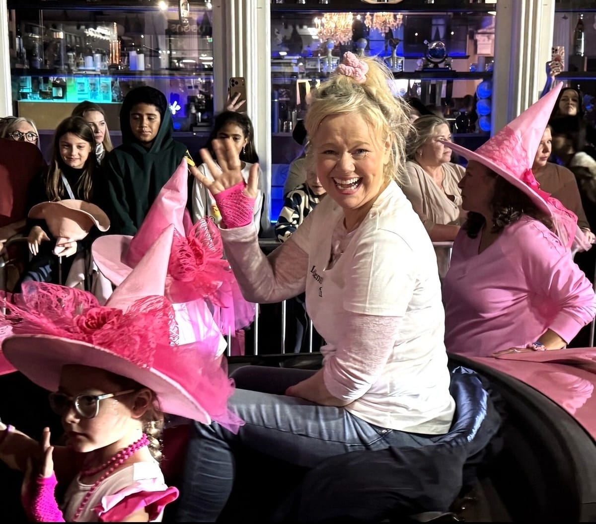 A laughing woman waving from a parade float with friends and a young girl, all in bright pink witch hats with feathers and tulle, the crowd and lit storefronts behind them