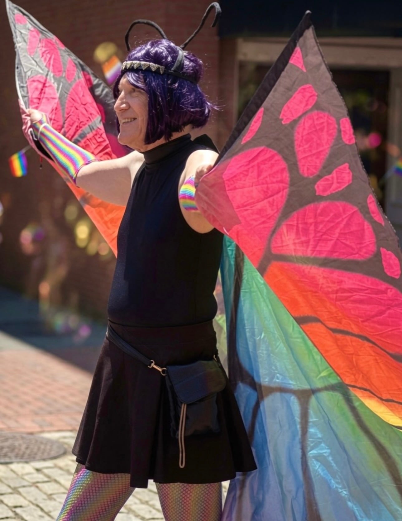 A performer in a monarch-butterfly costume with large colorful wings and a purple wig on a brick street during Salem Pride