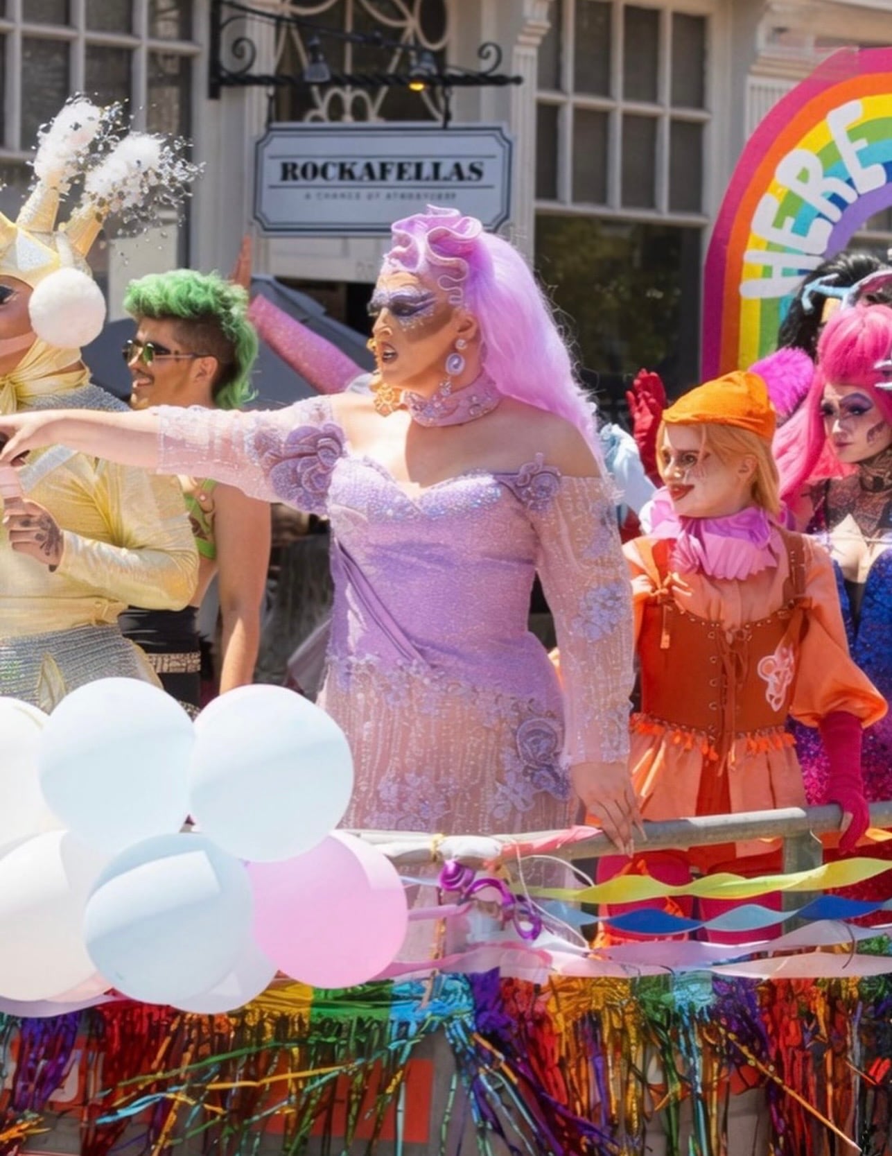 Drag performers in elaborate sequined costumes on a rainbow-decorated Pride float in front of Rockafellas in downtown Salem
