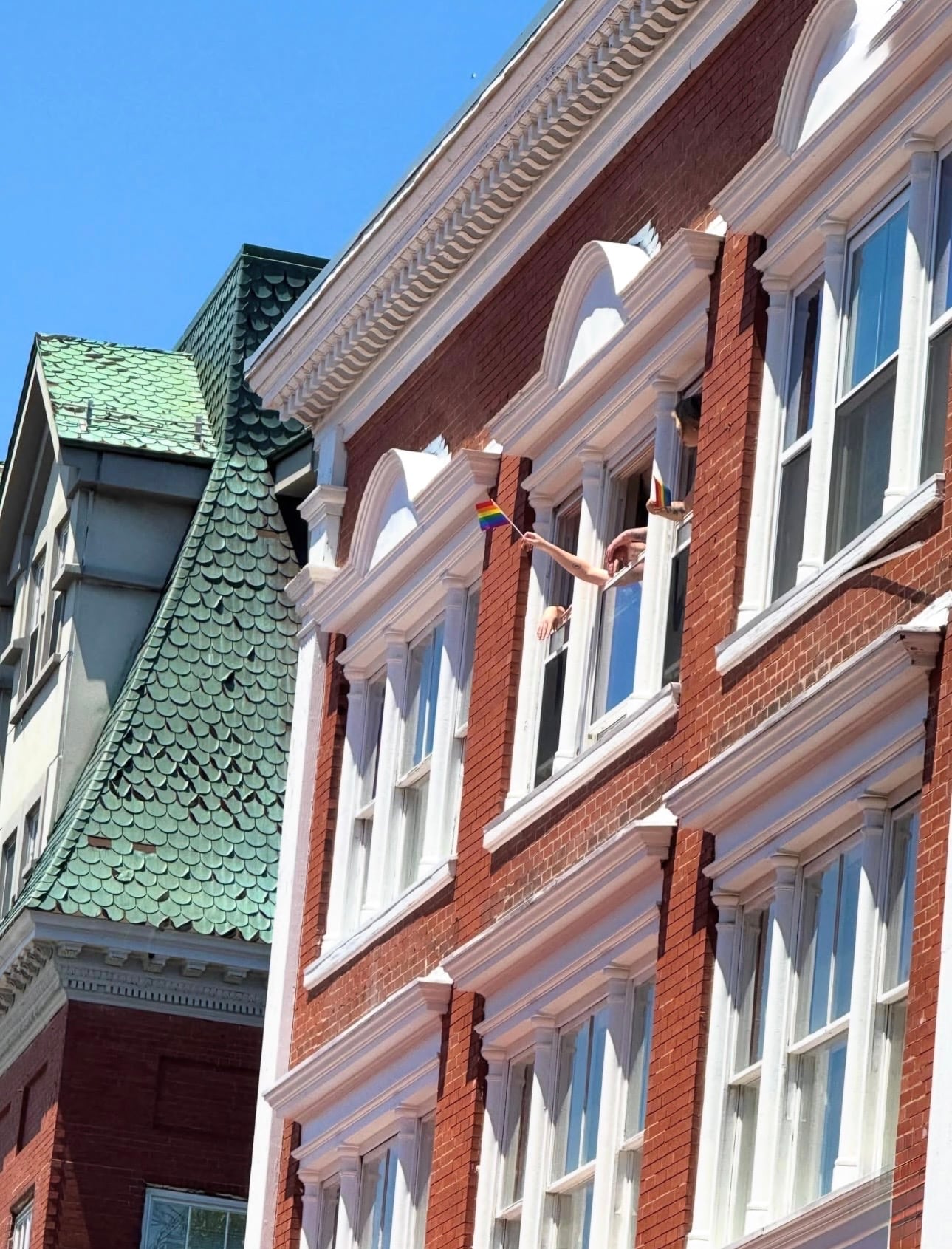 People leaning from an upper window of an ornate downtown Salem building, waving rainbow Pride flags, beside a green scalloped mansard roof