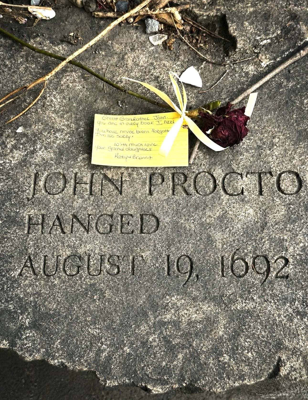 The John Proctor memorial bench at the Salem Witch Trials Memorial, inscribed 'hanged August 19, 1692', with a rose and a handwritten note left by a visitor