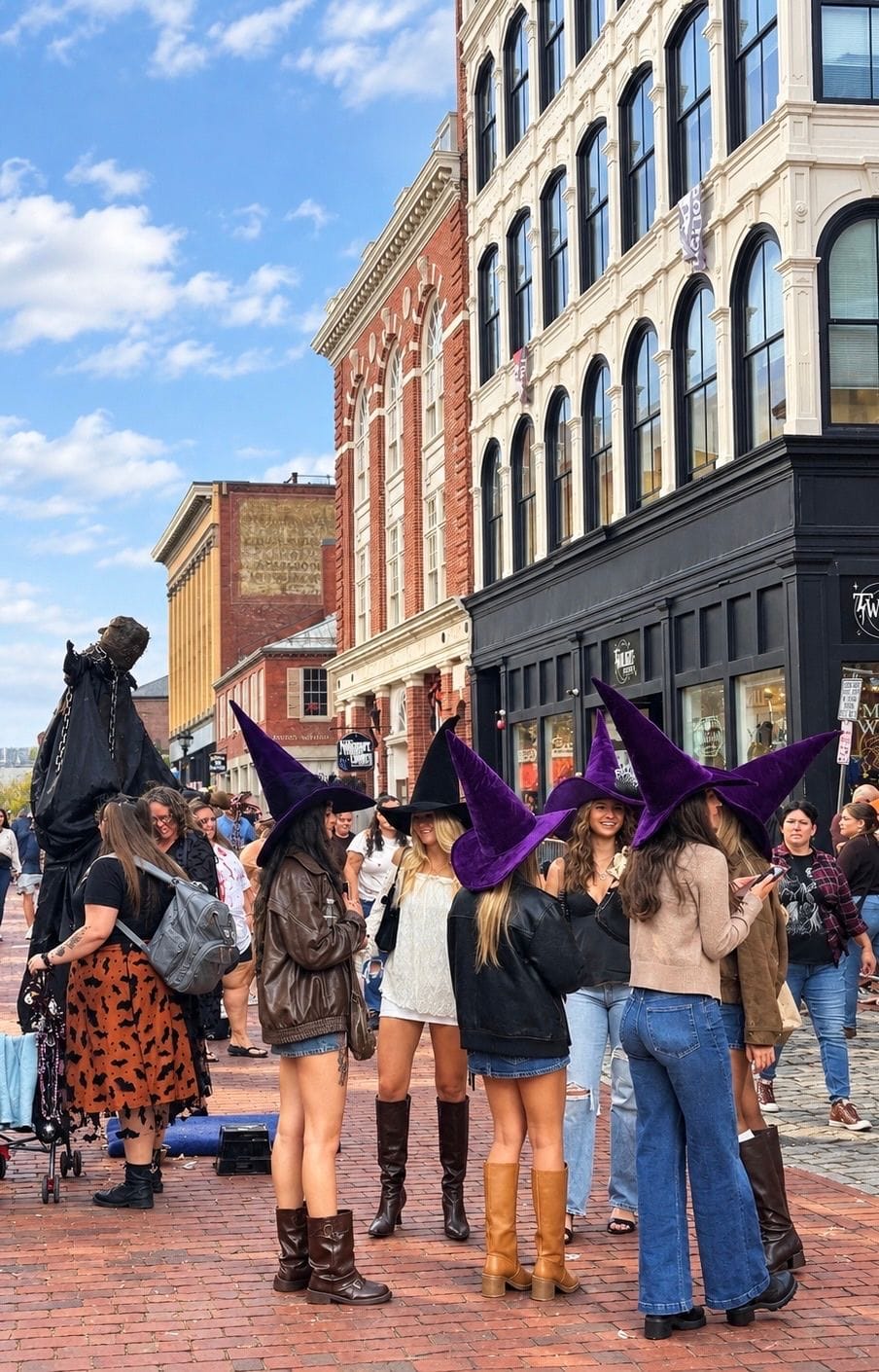 A group of friends in matching tall purple witch hats on the brick Essex Street pedestrian mall on a sunny October day, a robed stilt walker in chains behind them and the old brick and cast-iron storefronts beyond