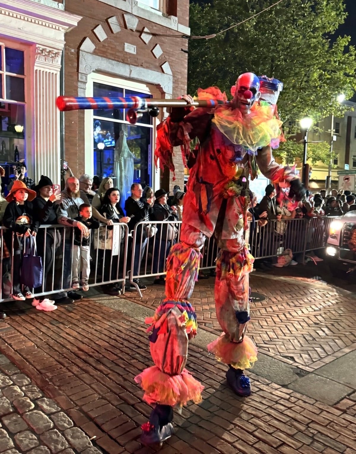 A towering stilt-walking clown in a rainbow ruffled costume and creepy clown mask, carrying a giant striped mallet on his shoulder past the crowd lining the barricades at night