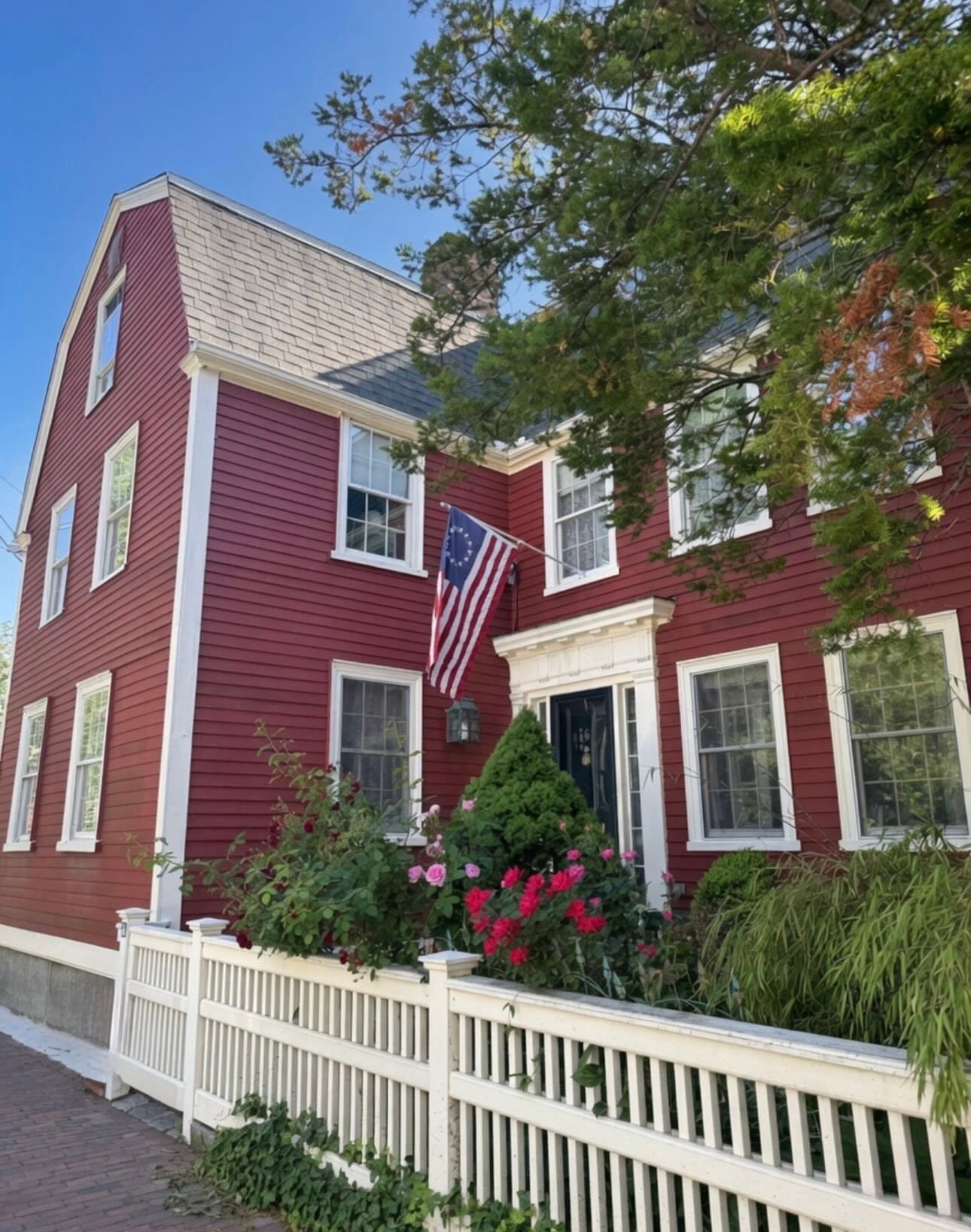 A deep-red gambrel-roofed colonial house with white trim, a 13-star flag, and a rose garden behind a white picket fence in Salem
