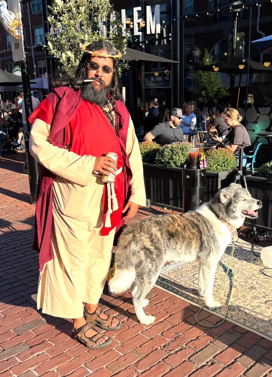 A street performer in a cream and deep red robe with a woven crown and sunglasses stands on the brick pedestrian mall beside a large grey and white dog, with café tables, planters of mums and the rooftop bar sign at Hotel Salem behind him