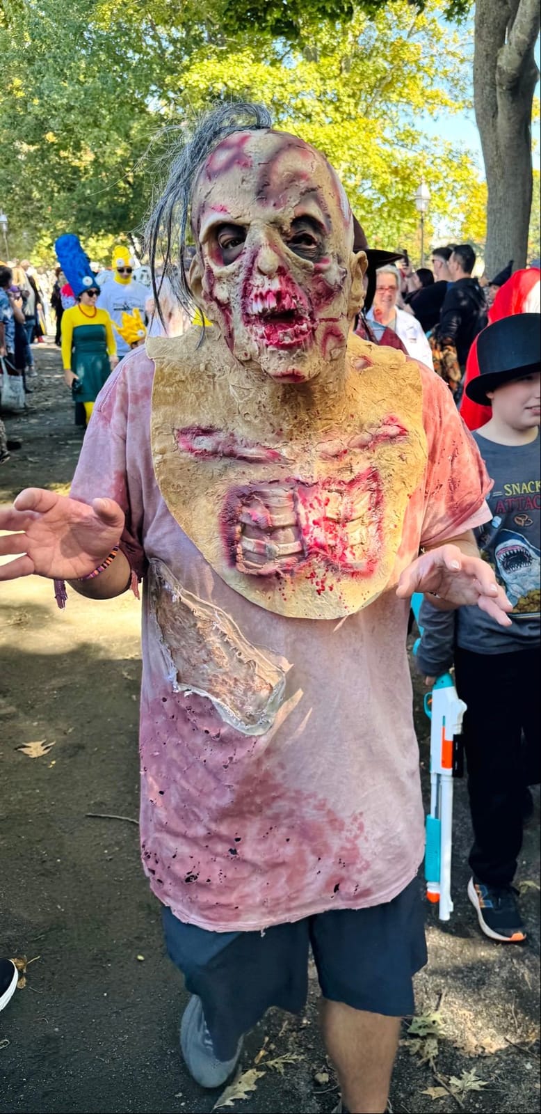 A zombie in a melting latex mask with exposed ribs on his chest and a bloodstained pink shirt, arms out, among the crowd on Salem Common
