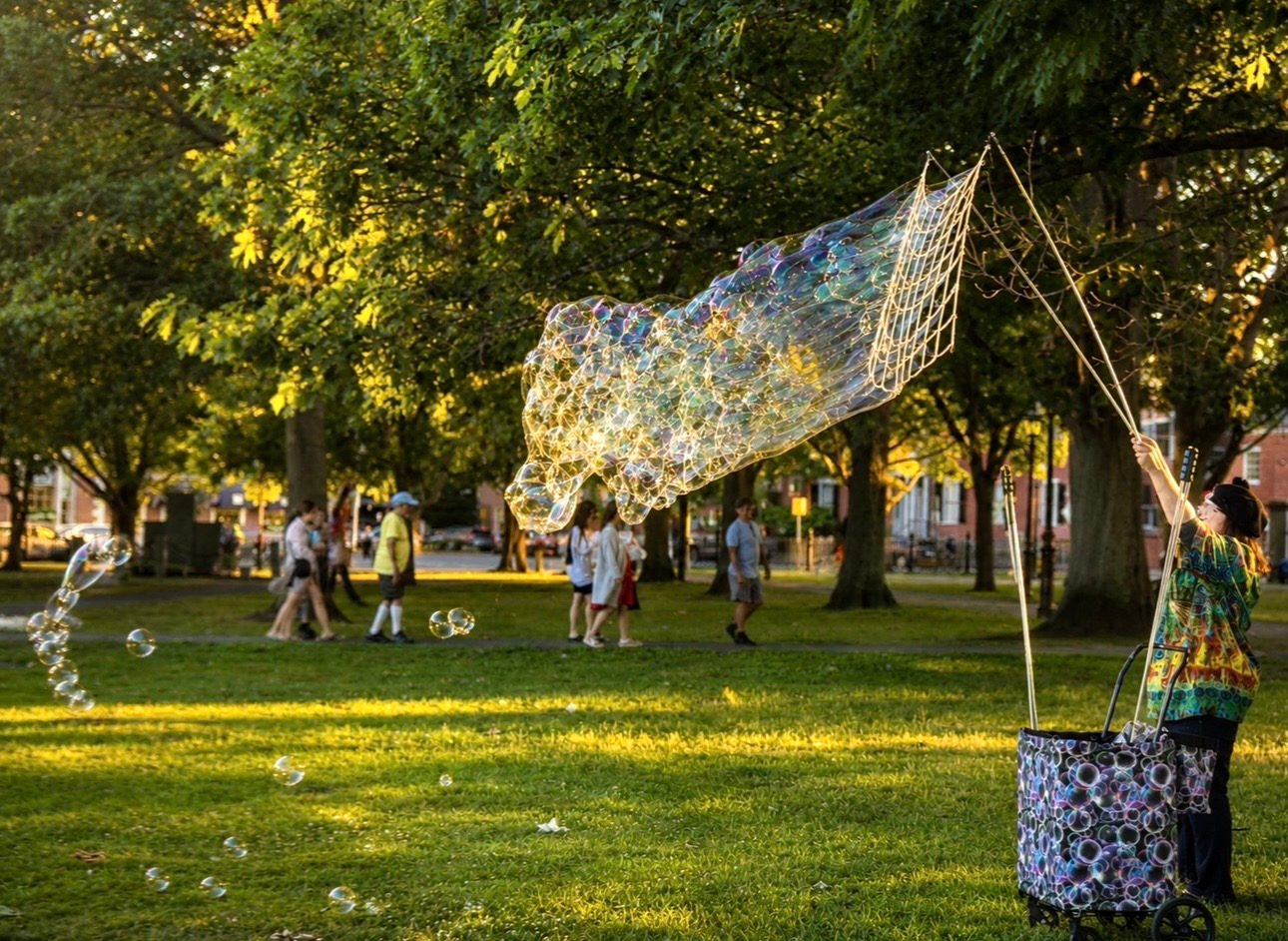 A street performer in tie-dye creates a glowing net of giant soap bubbles beneath the trees on Salem Common at golden hour