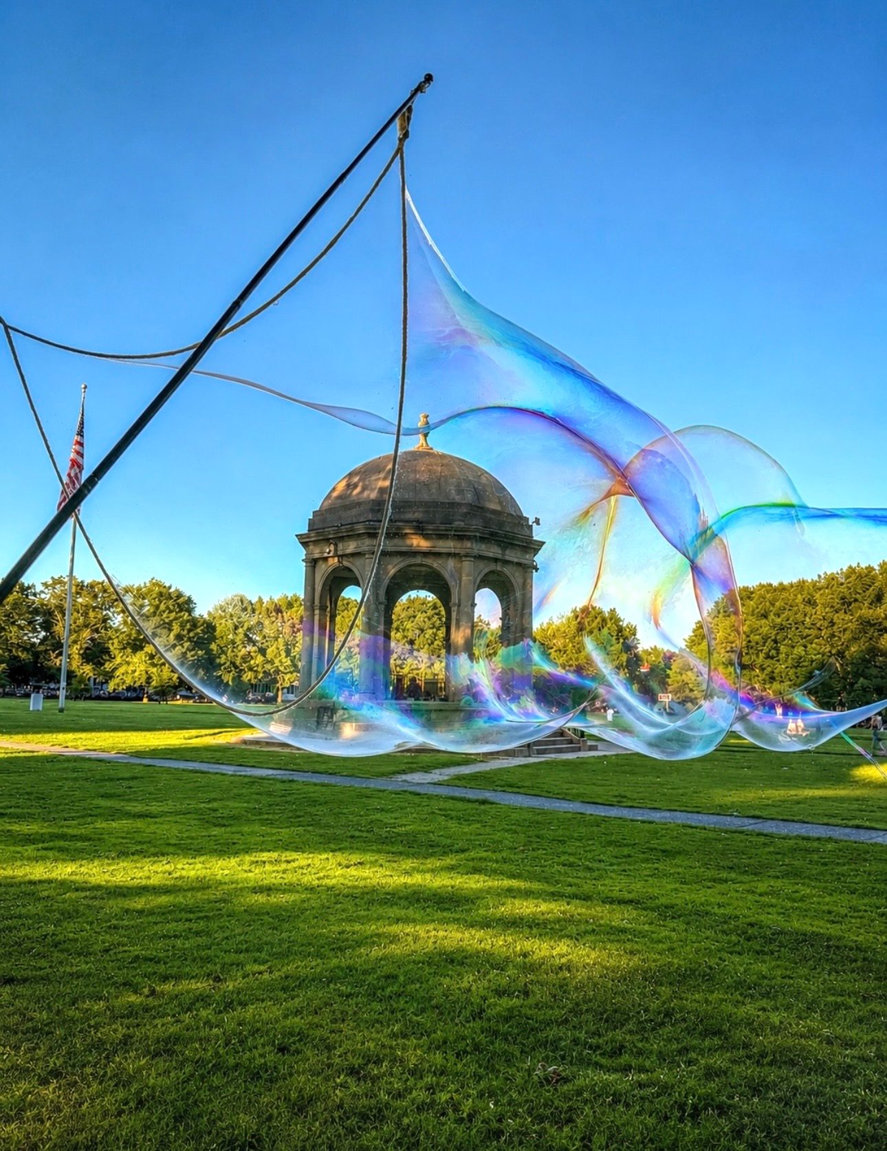 A long swirling ribbon of soap bubbles catches the light in front of the Salem Common bandstand and American flag