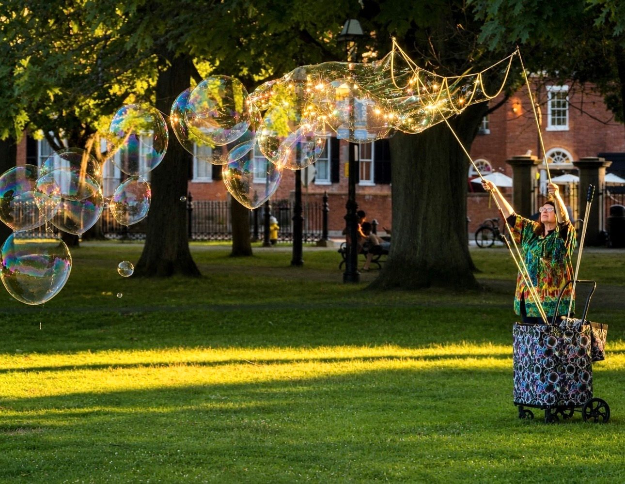A trail of large glowing soap bubbles floats across Salem Common at sunset as a performer works a bubble wand