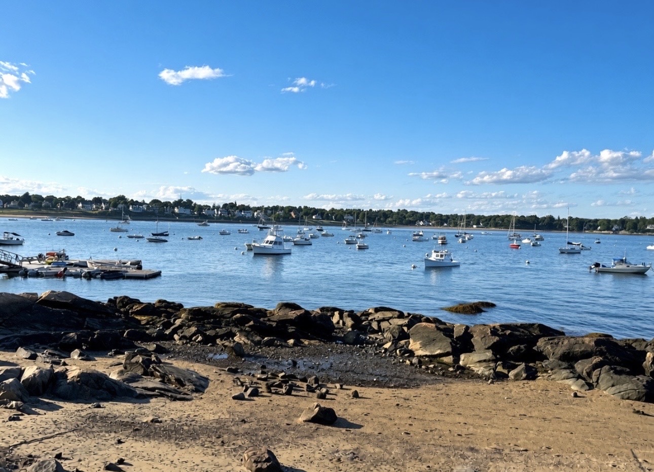 Recreational boats moored in Salem Harbor seen from a sandy beach with rocky outcrops, houses and a church steeple on the far shore, Salem MA