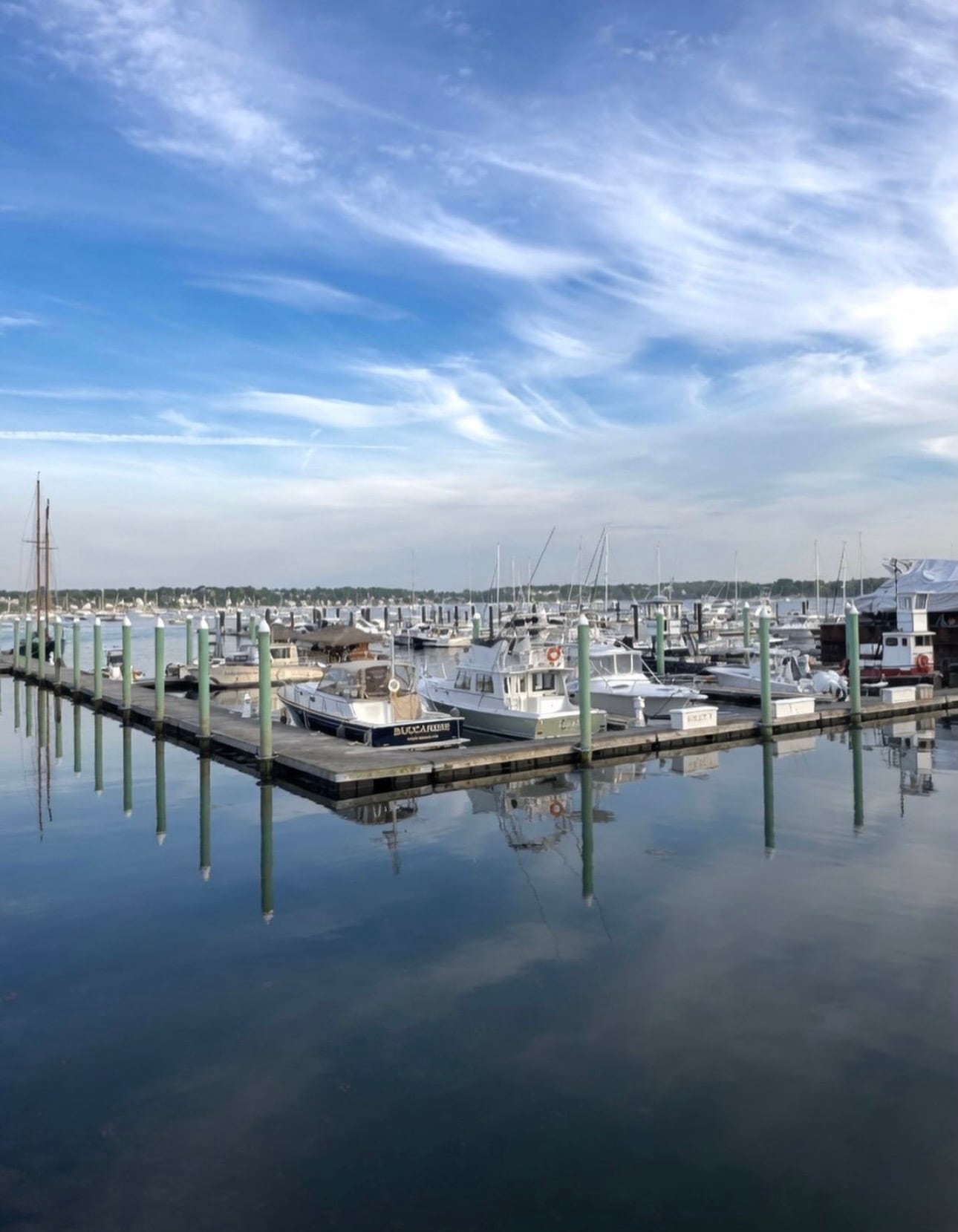 Boats and a schooner mast reflected in the calm water of a Salem Harbor marina under a wispy blue sky