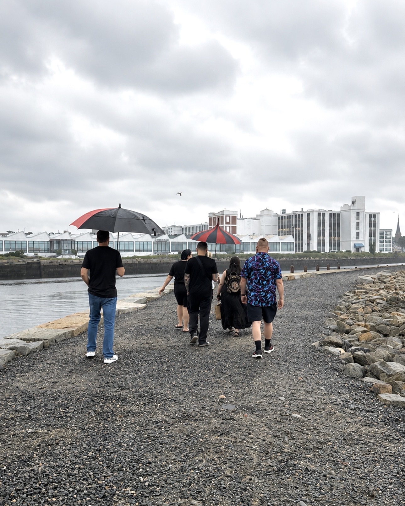 A group of visitors with red-and-black umbrellas walking the rocky causeway along Salem Harbor toward the Pickering Wharf waterfront, Salem MA