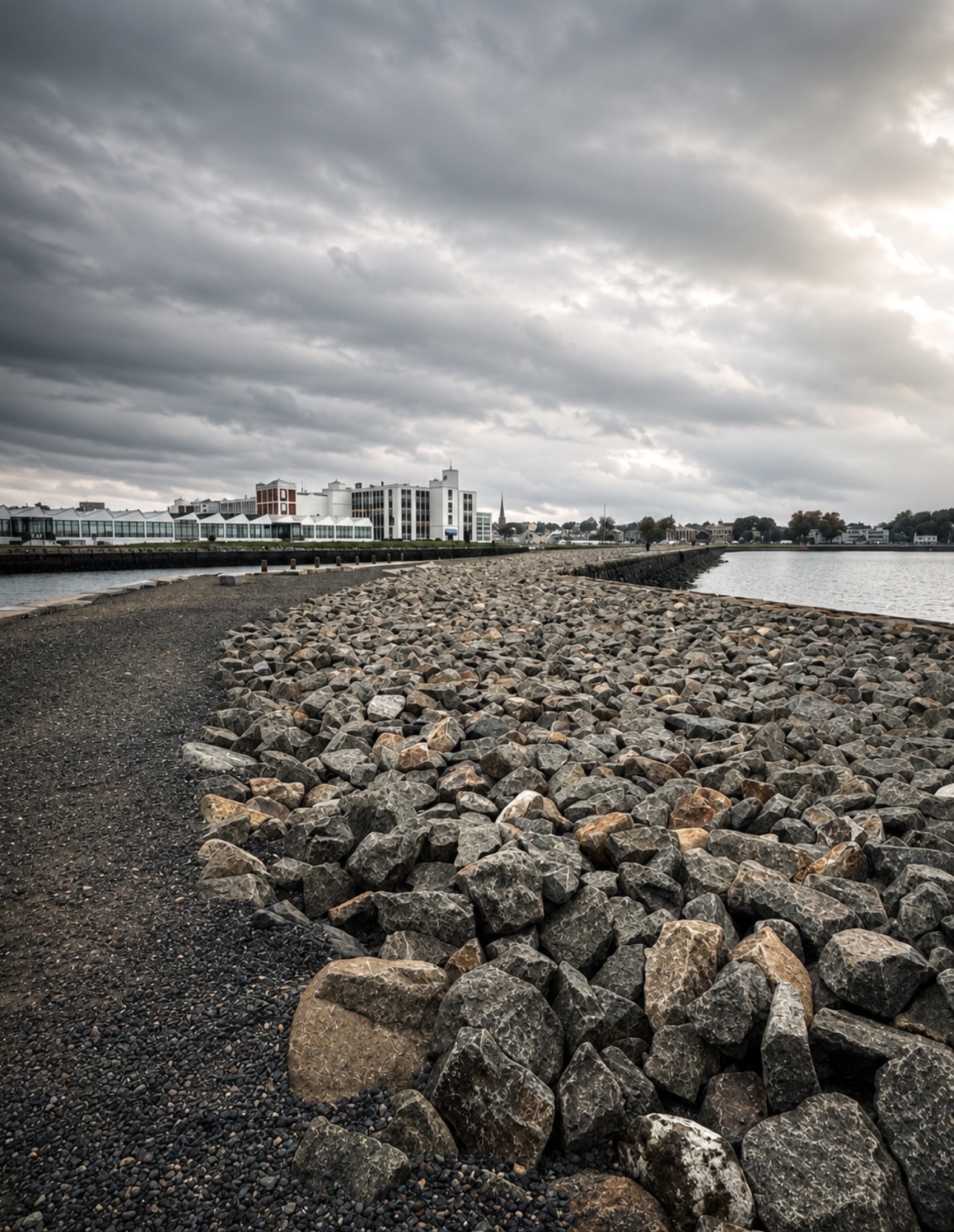 Granite riprap breakwater curving along Salem Harbor toward the Pickering Wharf waterfront under a heavy cloudy sky, Salem MA