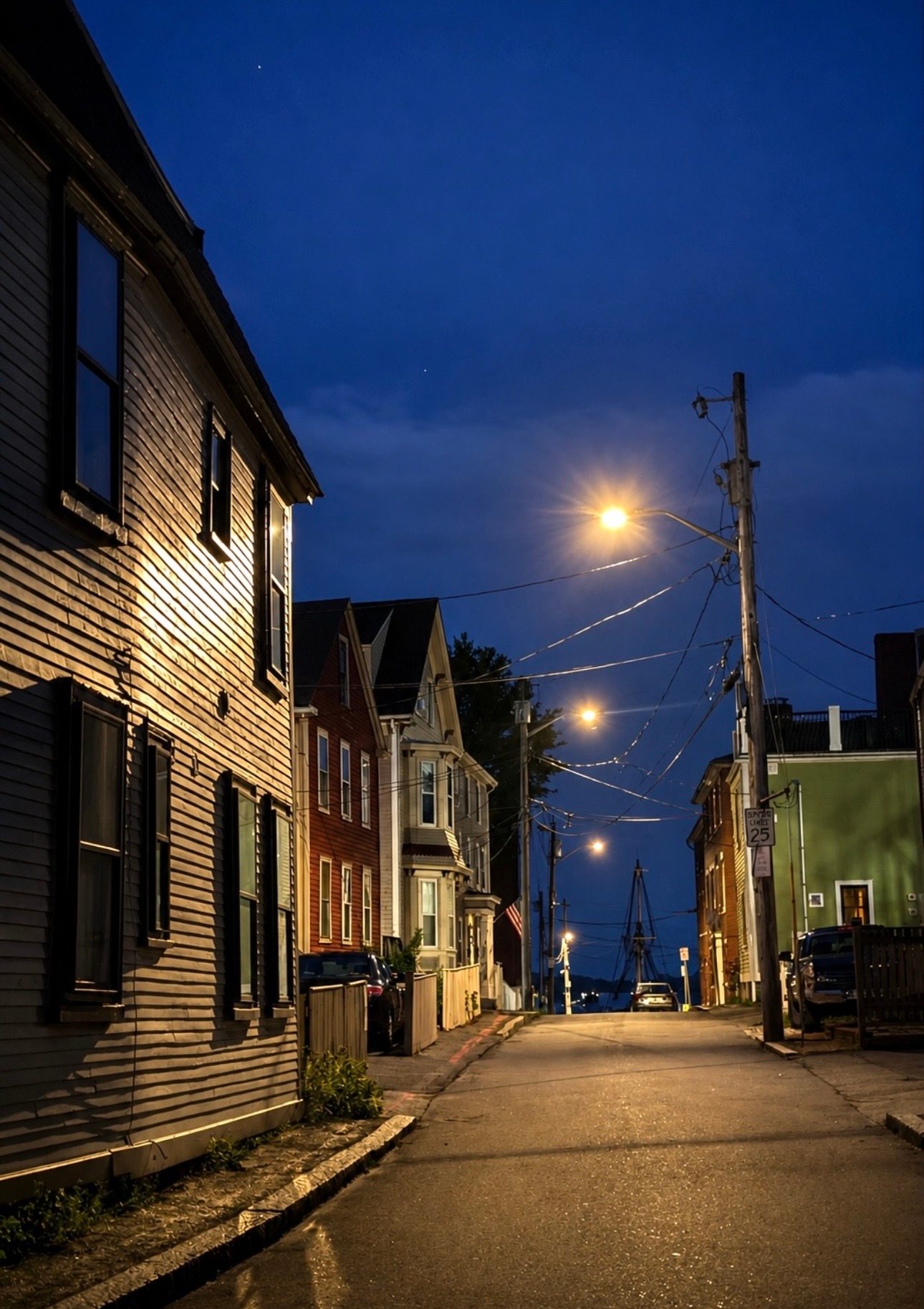 A quiet residential Salem street of clapboard houses sloping toward the harbor at blue hour, a tall-ship mast in the distance