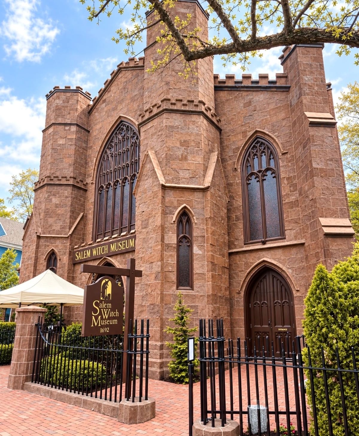 The Salem Witch Museum's brown stone Gothic Revival exterior with castellated towers and tall pointed-arch windows, its hanging sign reading Salem Witch Museum 1692, behind a black iron fence on a bright spring day