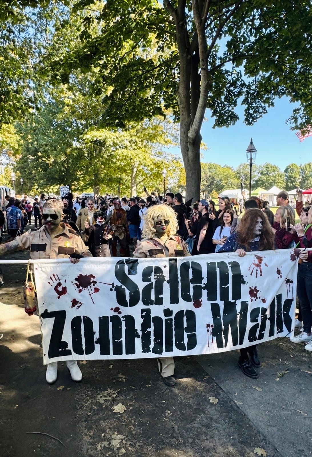 Zombies holding the blood-spattered Salem Zombie Walk banner at the front of the walk, a big crowd of costumed walkers behind them under the trees on Salem Common