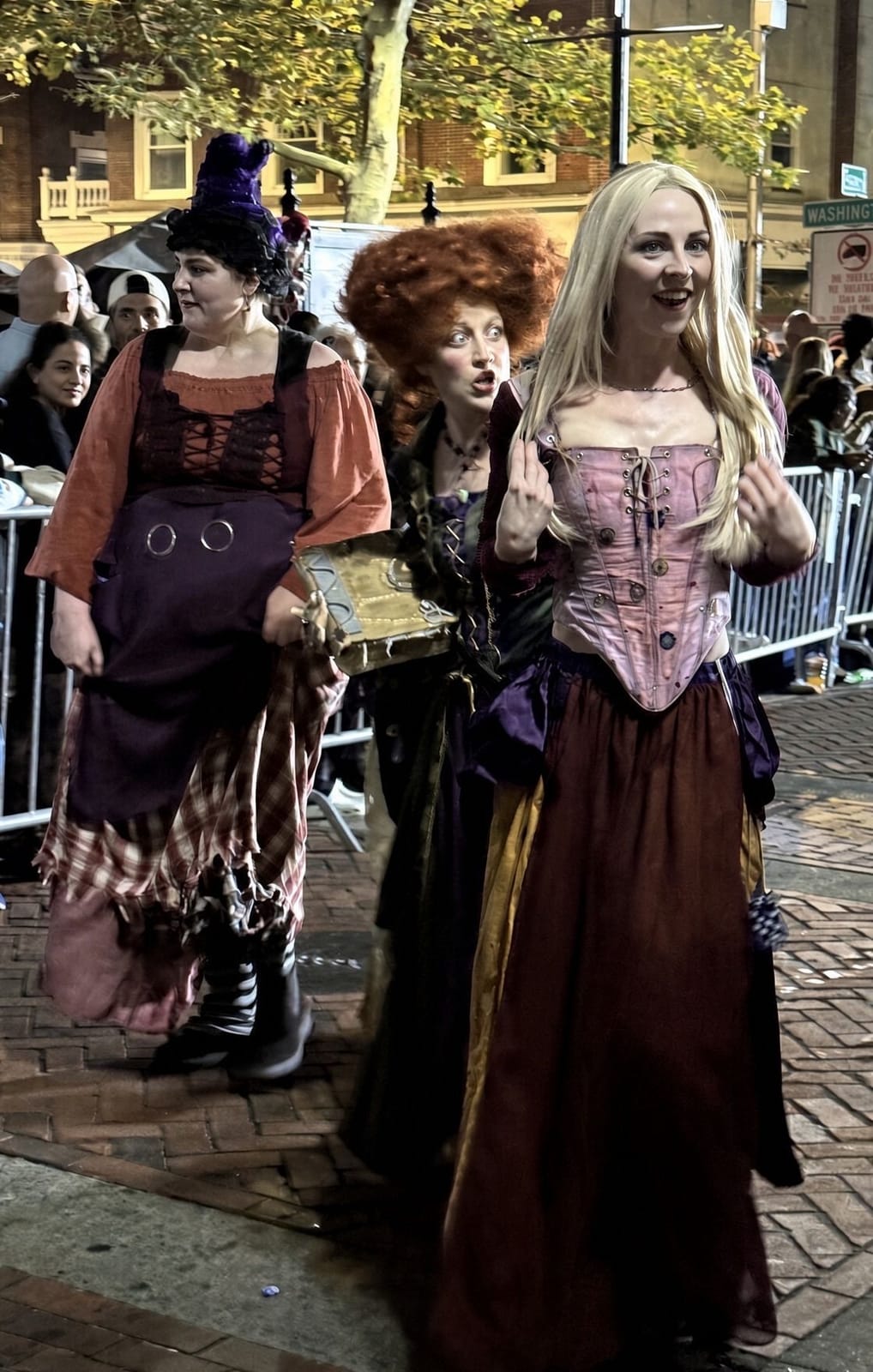 Three women dressed as the Sanderson sisters from Hocus Pocus, one holding the spell book, walking the brick parade route at night with the crowd behind the barricades