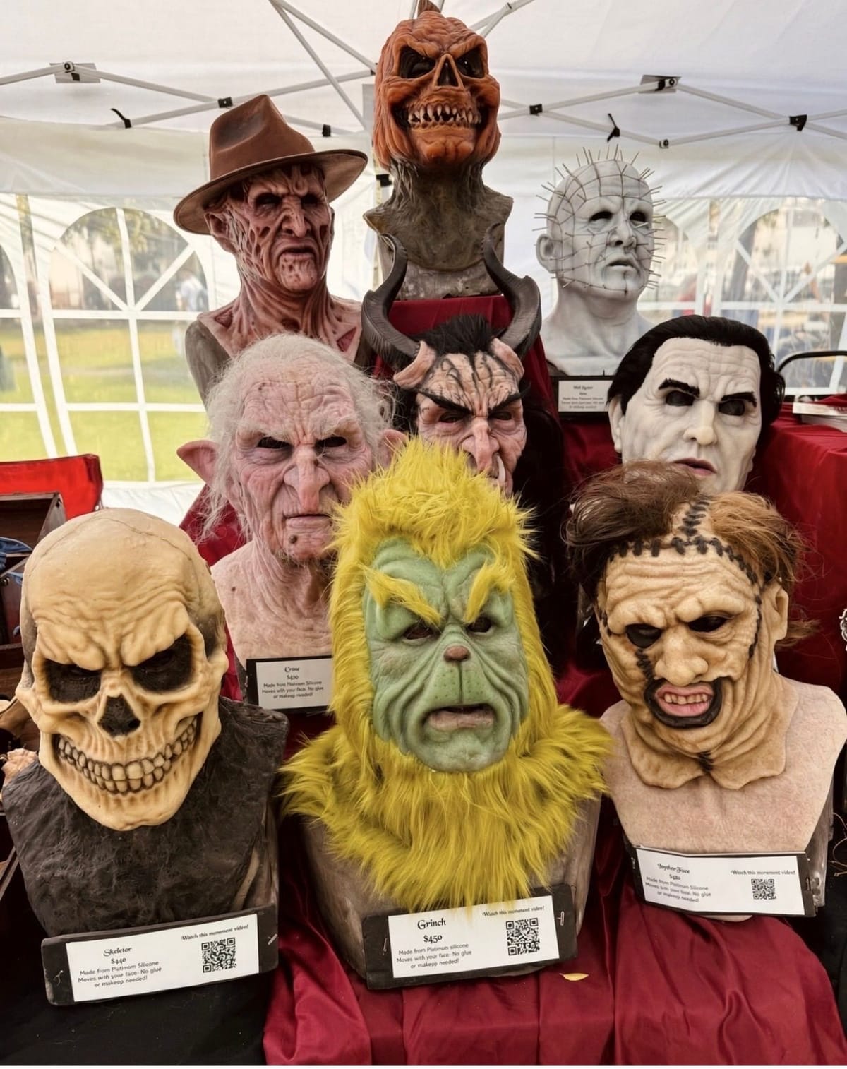 Ten lifelike silicone horror masks on display busts in a white vendor tent — Freddy Krueger in his fedora, a jack-o-lantern-headed creature, Pinhead, a horned demon, a pale vampire, an old crone, a skull, a furry green Grinch and Leatherface — over red cloth, with price cards and QR codes in front