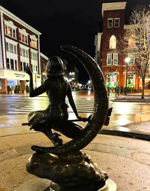 Bronze statue of Samantha from Bewitched, seated on a broom in front of a crescent moon, on its plinth at Lappin Park at night with wet pavement and lit storefronts behind.