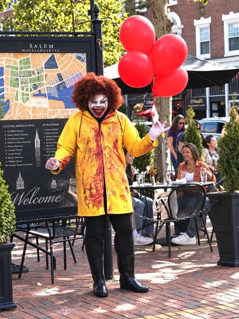 A street performer in clown makeup, a red wig and a blood-spattered yellow raincoat holds a bunch of red balloons beside a large Salem Massachusetts welcome map sign, with cafe tables and seated visitors behind