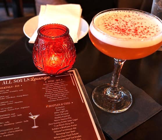 A coupe glass of pale orange cocktail topped with foam and red bitters, beside a red glass candle and the All Souls Lounge drinks menu