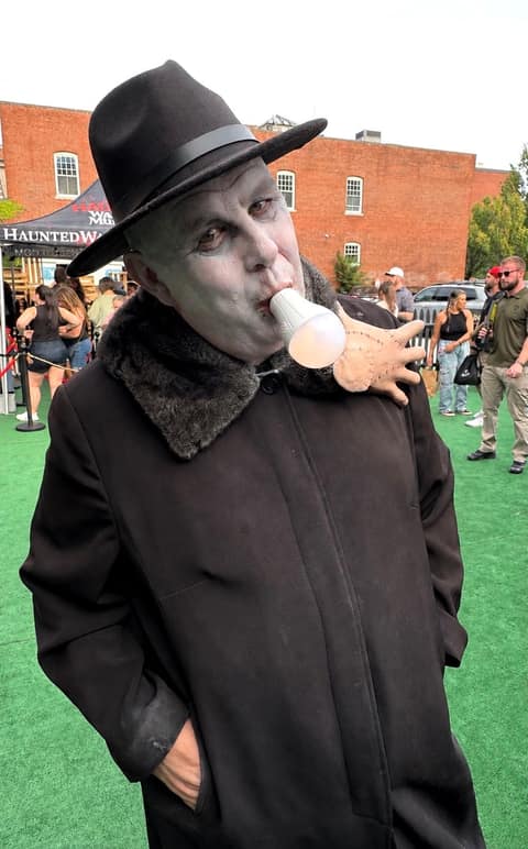 A performer in grey undead makeup, a black fedora and a fur-collared coat, holding a light bulb in their mouth on the green outside the Haunted Warren Museum