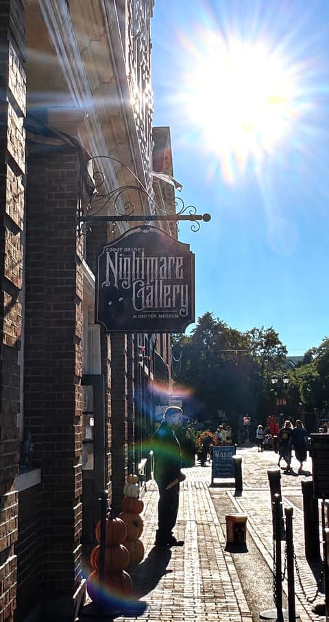 The wrought-iron sign for Count Orlok's Nightmare Gallery & Monster Museum against a sun-flared sky, with stacked pumpkins by the door on the Essex Street pedestrian mall