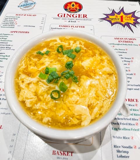 A bowl of egg drop soup topped with sliced scallions resting on the Ginger ramen and Asian fusion menu